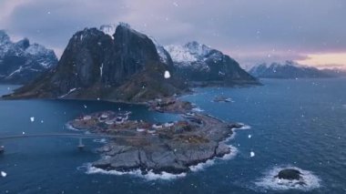 Karlı dağların havadan görünüşü, rorbu evleri, deniz, köprü ve yol olan adalar, kışın kar yağarken gün doğumunda mor bulutlu gökyüzü. Norveç, Lofoten adalarındaki Hamnoy köyünün en iyi insansız hava aracı görüntüsü. Dramatik