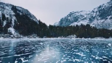 Kar yağarken donmuş gölün alçaktan uçan insansız hava aracı görüntüsü. Kışın gün batımında dağlar. Lofoten Adaları, Norveç. Buzlu nehrin üst manzarası, buzdaki yansıma, karlı kayalar, gökyüzü. Bulutlu hava