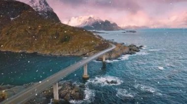 Köprünün havadan görünüşü, dalgalı deniz ve kışın karlı gündoğumunda karlı dağlar. Lofoten Adaları, Norveç. Güzel yol, su, kayalar, şafak vakti bulutlu pembe gökyüzü.