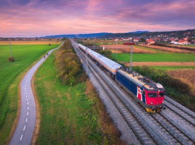Alp köyü, dağlar ve yeşil tarlalar yakınında hareket eden yük treninin hava manzarası, baharda gün batımında bisiklet yolu. Yazın tren, demiryolu, yol, çayır manzarası. Hırvatistan 'daki tren istasyonu