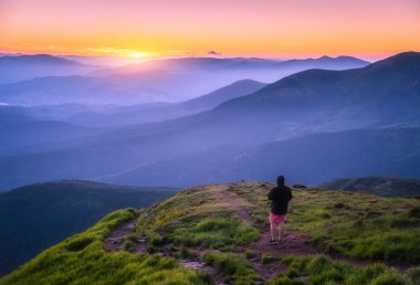 Ukrayna 'nın dağının tepesinde bir adam var. Yazın gün batımında yeşil çimenli tepe ve güzel dağ vadisi. Sportif bir adamla manzara, sis bulutu, alacakaranlıkta turuncu gökyüzü. Yürüyüş. Bahar