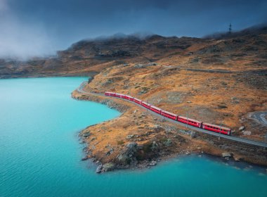 Modern kırmızı trenin havadan görünüşü, turkuaz dağ gölü ve sonbaharda günbatımında dramatik bulutlu gökyüzü. Bernina Express, İsviçre. Alplerdeki tren manzarası, dolambaçlı demiryolu, turuncu tepeler. Seyahat