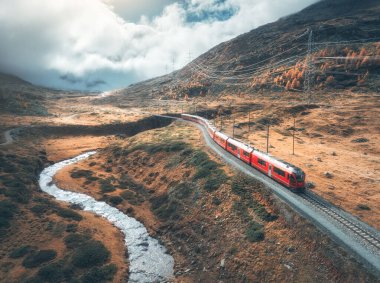 Modern kırmızı trenin, nehrin, dağların, turuncu tepelerin ve ağaçların, sonbaharda günbatımında bulutlu gökyüzünün havadan görünüşü. Bernina Express, İsviçre. Alplerdeki trenin en üst manzarası, sonbaharda demiryolu dolambaçlı.