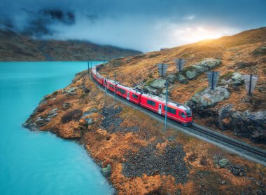 Modern kırmızı trenin havadan görünüşü, turkuaz dağ gölü ve sonbaharda günbatımında dramatik bulutlu gökyüzü. Bernina Express, İsviçre. Alplerdeki tren manzarası, dolambaçlı demiryolu, turuncu tepeler. Seyahat