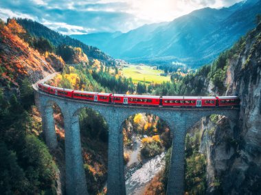 Landwasser 'daki modern kırmızı trenin sonbaharda günbatımında turuncu orman ve dağlardaki viyadük manzarası. Bernina Express, İsviçre. Alplerdeki tren manzarası, demiryolu, sonbaharda renkli ağaçlar.