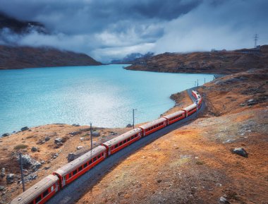 Modern kırmızı trenin havadan görünüşü, turkuaz dağ gölü ve sonbaharda günbatımında dramatik bulutlu gökyüzü. Bernina Express, İsviçre. Alplerdeki tren manzarası, dolambaçlı demiryolu, turuncu tepeler. Seyahat