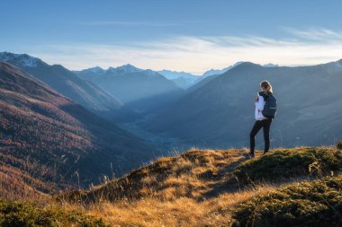 Sırt çantalı genç bir kadın çimenli dağ sırtında durup sonbaharda altın saat günbatımında yüksek dağ zirveleri olan vadiden ilham verici manzaranın tadını çıkarıyor. İtalya 'da yürüyüş, Levigno. Alpli Kız