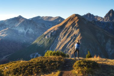 Sırt çantalı genç bir kadın çimenli dağ yolunda durup sonbaharda altın saat batımında yüksek dağ zirveleri olan vadinin tadını çıkarıyor. İtalya 'da yürüyüş ve yürüyüş, Levigno. Sonbaharda Alplerdeki Kız