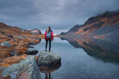 Sonbaharda bir kayanın üzerinde sırt çantalı genç bir kadın yürüyüşçü. Kız ve nefes kesici dağ alp gölü manzarası çevredeki tepeleri, turuncu otları ve bulutlu dramatik gökyüzünü yansıtıyor. İsviçre sonbaharda