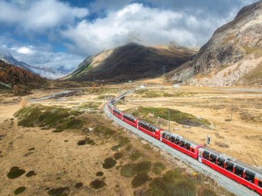 Modern kırmızı trenin, dağların, turuncu tepelerin ve ağaçların havadan görünüşü güneşli sonbahar gününde bulutlu bir gökyüzü. Bernina Express, İsviçre. Alplerdeki trenin en üst manzarası, sonbaharda demiryolu dolambaçlı.