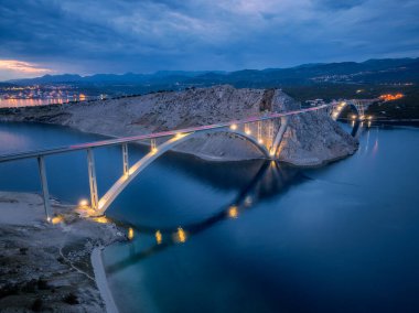 Geceleri mavi deniz ve köprü manzarası. Günbatımında aydınlık, ada, dağlar, su ve bulutlu gökyüzü olan modern Krk köprüsü. Yol manzarası, şehir ışıkları, alacakaranlıkta kayalar. Mimari