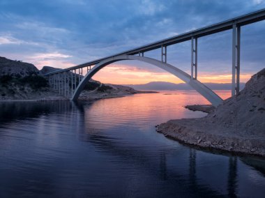 Renkli günbatımında mavi deniz ve köprü manzarası. Altın saat boyunca modern Krk Köprüsü adası, dağlar, kayalar, sudaki yansıma ve bulutlu gökyüzü. Köprülerin üst drone görüntüsü. Mimari