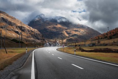 Boş asfalt yolu sonbaharda günbatımında bulutların içinde Alp Dağları 'ndan geçiyor. Görkemli Alpler, portakal ağaçları ve otlarla dolu manzara, sonbaharda dramatik bulutlu bir gökyüzü. Dağ geçidinde yol. İsviçre