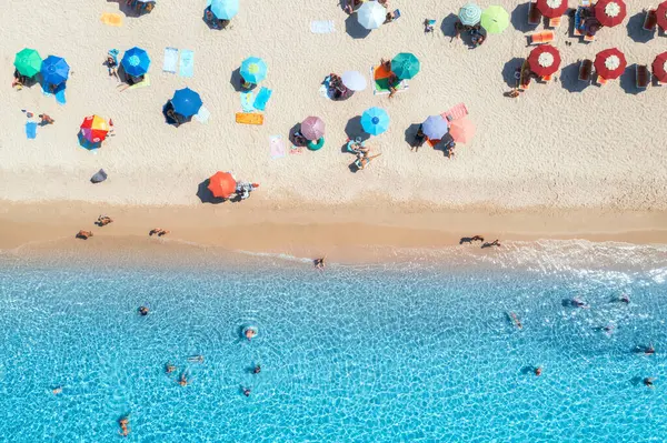 Kumlu sahildeki renkli şemsiyeler, güneşli yaz günlerinde mavi denizde yüzen insanlar. Sardunya, İtalya. Berrak gök mavisi suyu olan tropik bir yer. Seyahat ve tatil. Deniz kenarının üst görünümü