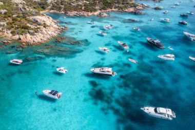 Lüks yatların ve teknelerin, seyrek bitki örtüsü olan kayalık bir sahil şeridi yakınındaki Akdeniz 'in berrak turkuaz sularında gezindiği hava manzarası. Üst dron görüntüsü. İtalya, Sardunya 'da yaz tatili
