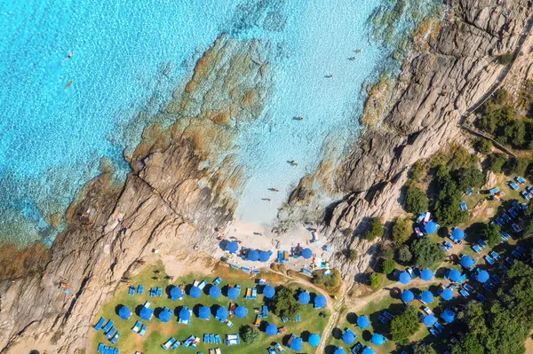 Deniz koyunun havadan görünüşü, kumlu plaj şemsiyeli, taşlı ve kayalıklı yaz günbatımında suda. Sardunya, İtalya. Deniz lagünüyle tropik manzara, yüzen insanlar, mavi su. Yukarıdan drone görünümü