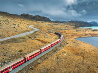 Gökyüzü, sonbaharda günbatımında gökyüzü bulutlu ve kayalık göl kenarındaki modern kırmızı trenin görüntüsü. Bernina Express, İsviçre. Alplerdeki tren manzarası, dolambaçlı demiryolu, sonbaharda turuncu tepeler.