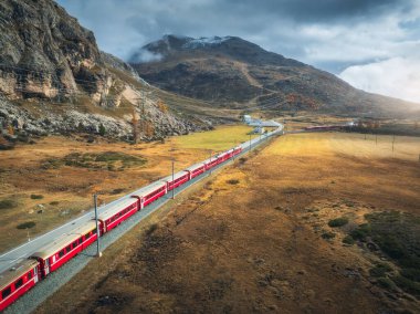 Modern kırmızı trenin, dağların, turuncu çayır ve ağaçların, sonbaharda günbatımında bulutlu gökyüzünün havadan görünüşü. Bernina Express, İsviçre. Alplerdeki trenin en üst manzarası, sonbaharda demiryolu dolambaçlı.