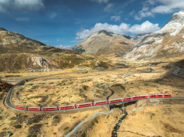 Modern kırmızı trenin, dağların, turuncu çayır ve ağaçların, sonbaharda günbatımında bulutlu gökyüzünün havadan görünüşü. Bernina Express, İsviçre. Alplerdeki trenin en üst manzarası, sonbaharda demiryolu dolambaçlı.