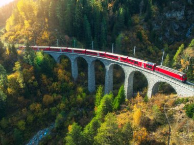 Landwasser 'daki modern kırmızı trenin sonbaharda günbatımında turuncu orman ve dağlardaki viyadük manzarası. Bernina Express, İsviçre. Alplerdeki tren manzarası, demiryolu, sonbaharda renkli ağaçlar.