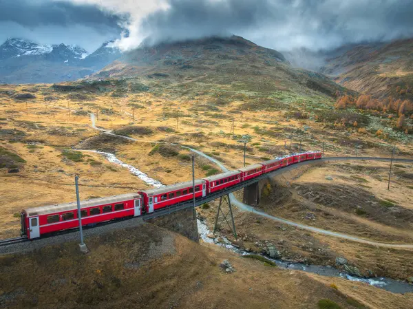 Modern kırmızı trenin hava manzarası sonbaharda günbatımında İsviçre Alp Vadisi 'ndeki köprülerden geçiyor. Bernina Ekspres treni, turuncu çimenler, dağlar, nehir ve sonbaharda bulutlu gökyüzü. Demiryolu.