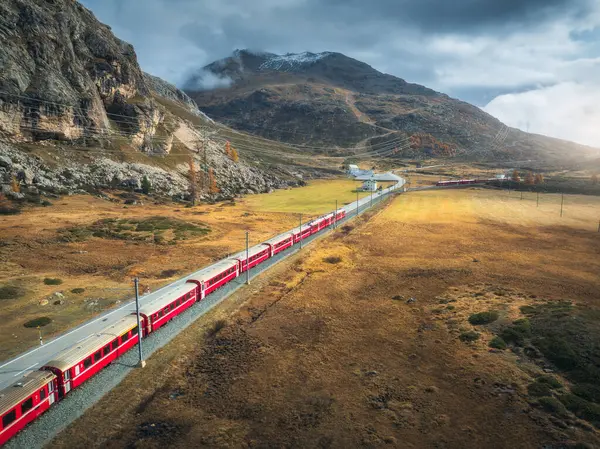 Modern kırmızı trenin, dağların, turuncu çayır ve ağaçların, sonbaharda günbatımında bulutlu gökyüzünün havadan görünüşü. Bernina Express, İsviçre. Alplerdeki trenin en üst manzarası, sonbaharda demiryolu dolambaçlı.
