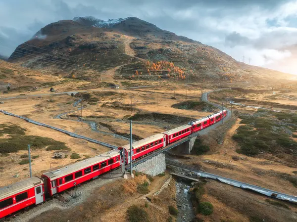 Modern kırmızı trenin hava manzarası sonbaharda günbatımında İsviçre Alp Vadisi 'ndeki köprülerden geçiyor. Bernina Ekspres treni, turuncu çimenler, dağlar, nehir ve sonbaharda bulutlu gökyüzü. Demiryolu.