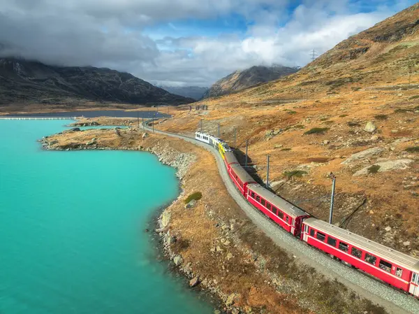 Modern kırmızı trenin havadan görünüşü, turkuaz dağ gölü ve sonbaharda günbatımında dramatik bulutlu gökyüzü. Bernina Express, İsviçre. Alplerdeki tren manzarası, dolambaçlı demiryolu, turuncu tepeler. Seyahat