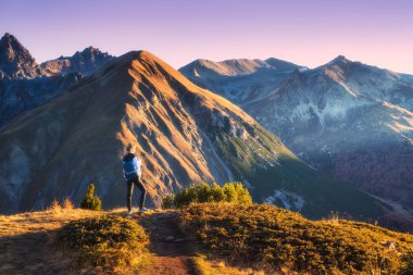 Sonbaharda altın saat batımında yüksek dağ zirveleri olan vadideki çimenli dağ yolunda sırt çantalı genç bir kadın. İtalya 'da yürüyüş ve yürüyüş, Levigno. Sonbaharda Alplerdeki Kız