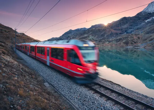 Gök mavisi Bianco Gölü 'nün yanındaki modern kırmızı tren, sonbaharda günbatımında karlı kayalar ve pembe gökyüzü. Bernina Express, İsviçre. Alpler 'de bulanık tren, alacakaranlıkta suda yansıma. Seyahat