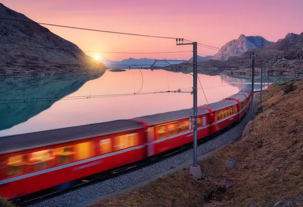 Gök mavisi dağ Bianco gölü yakınlarındaki modern kırmızı tren, karlı kayalar ve sonbaharda günbatımında renkli pembe gökyüzü. Bernina Express, İsviçre. Alplerde bulanık tren, alacakaranlıkta suda yansıma.