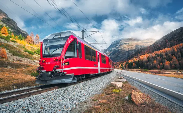 Kızıl modern yolcu treni sonbaharda günbatımında İsviçre Alplerinde demiryolunda hareket ediyor. Trenle yüksek dağlar, yollar, sonbaharda portakal ağaçları. Bernina Express, İsviçre. Geçişi koruyun.