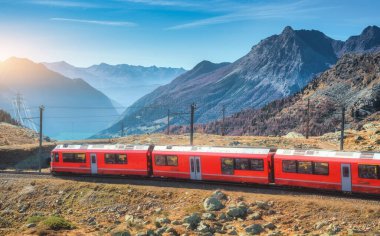 Kızıl modern tren sonbaharda İsviçre Alpleri 'nden geçiyor. Hareket eden tren, dağlık vadi, turuncu tepeler, sonbaharda gün batımında mavi gökyüzü. Demiryolu seyahati Bernina Ekspresi, İsviçre