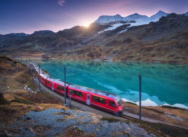 Gök mavisi dağ Bianco Gölü 'nün yanındaki modern kırmızı tren, karlı kayalar ve sonbaharda günbatımında renkli gökyüzü. Bernina Express, İsviçre. Alpler 'de bulanık tren, demiryolu, alacakaranlıkta masmavi su