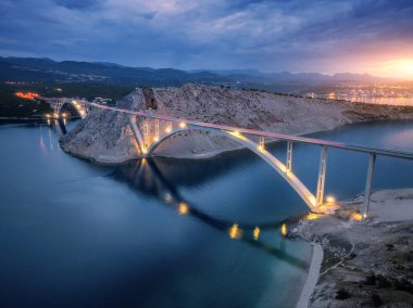 Geceleri mavi deniz ve köprü manzarası. Günbatımında aydınlık, ada, dağlar, su ve bulutlu gökyüzü olan modern Krk köprüsü. Yol manzarası, şehir ışıkları, alacakaranlıkta kayalar. Mimari