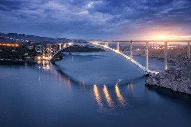 Geceleri mavi deniz ve köprü manzarası. Günbatımında aydınlık, ada, dağlar, su ve bulutlu gökyüzü olan modern Krk köprüsü. Yol manzarası, şehir ışıkları, alacakaranlıkta kayalar. Mimari