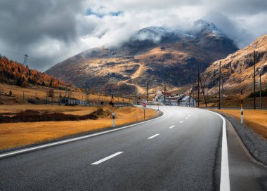 Boş asfalt yolu sonbaharda günbatımında bulutların içinde Alp Dağları 'ndan geçiyor. Görkemli Alpler, portakal ağaçları ve otlarla dolu manzara, sonbaharda dramatik bulutlu bir gökyüzü. Dağ geçidinde yol. İsviçre