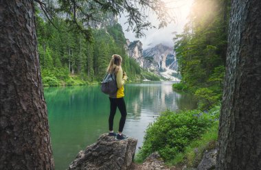 Sırt çantalı genç bir kadın Braies Gölü 'ndeki taşın üzerinde Alp Dağları' nda, bulutlu gökyüzü, suda yansıma, gün batımında yeşil ağaçlar. Dolomitler, İtalya 'ya yaz seyahati. Göldeki kız. Yürüyüş