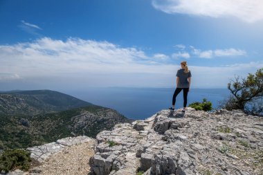 Uçurumda yürüyüş yapan genç sarışın kadın, bulutlu, güneşli mavi bir gökyüzünün altında Krk Adaları 'ndan Adriyatik Denizi' nin panoramik manzarasını seyrediyor. Dağ tepesindeki kız. Gezinti. Yazın seyahat.