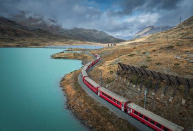 İsviçre Alpleri 'ndeki turkuaz göl boyunca kıvrılan kırmızı Bernina Ekspres treni, kayalık tepeler ve sonbahar dağları manzarasında dramatik bulutlu gökyüzü. Sonbaharda trenin üst görüntüsü