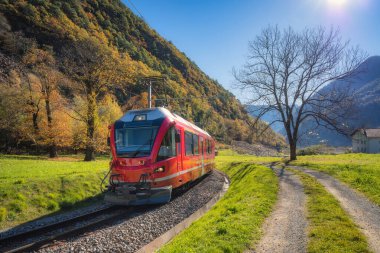 Kızıl Bernina Ekspresi İsviçre 'de güneşli bir sonbahar vadisinden geçiyor açık mavi gökyüzü altında yeşil çimenler, demiryolu rayları ve dağ yamaçları var. İsviçre Alplerinde modern tren