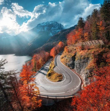 Sonbaharda günbatımında dönen dağ yolunun hava manzarası. Alpler, İtalya. Kavisli dağlık yol, karlı kayalar, ağaçlar, kırmızı yapraklar, göl, güneş ışınlı gökyüzü. Renkli. Sonbaharda dağ geçidinde yol manzarası