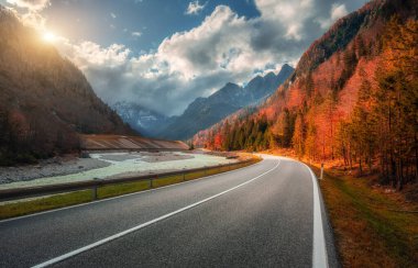 Sonbaharda gün batımında dağ yolu. Alpler, İtalya. Güzel kıvrımlı dağ yolu, karlı kayalar, ağaçlar, kırmızı yapraklar, nehir, bulutlu gökyüzü. Sonbaharda dağ geçidinde boş yolu olan renkli bir manzara.