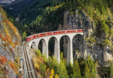 Alp dağlarındaki Landwasser viyadük üzerindeki modern kırmızı trenin havadan görünüşü, sonbaharda günbatımında renkli orman. Bernina Express, İsviçre. Alplerdeki tren manzarası, demiryolu, sonbaharda yeşil ağaçlar