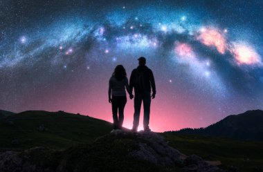 Dağlarda, pembe gece yıldızlı gökyüzünün altında parlayan Samanyolu 'nun üzerinde el ele tutuşan iki yürüyüşçü. Erkek ve kadın siluetleri, parlak yıldızlar ve samanyolu manzarası