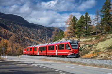 İsviçre Alpleri 'ndeki renkli sonbahar ormanlarından geçen Kızıl Bernina Ekspres treni, arka planda dramatik bulutlar, altın ağaçlar ve karlı dağ zirveleriyle İsviçre' de. Buzul Ekspres Treni