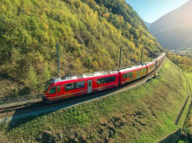 Alp dağlarındaki modern kırmızı trenin havadan görünüşü, sonbaharda günbatımında yeşil çayırlar. Bernina Express, İsviçre. Alplerdeki tren manzarası, sonbaharda demiryolu. Tren istasyonunda. Seyahat