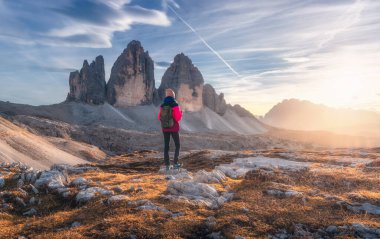 Sonbaharda gün batımında dağ zirvesinde sırt çantalı ve yüksek kayalıklı bir kız. Tre Cime, Dolomitler, İtalya. Yoldaki genç kadınla manzara, uçurumlar, bulutlu gökyüzü ve sonbaharda güneş. Yürüyüş