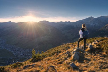 Sırt çantalı genç bir kadın sonbaharda altın saat batımında yüksek dağ zirveleri olan vadi manzaralı çimenli dağ sırtında duruyor. İtalya 'da yürüyüş, Levigno. Sonbaharda Alpler 'de bir kız. Seyahat.