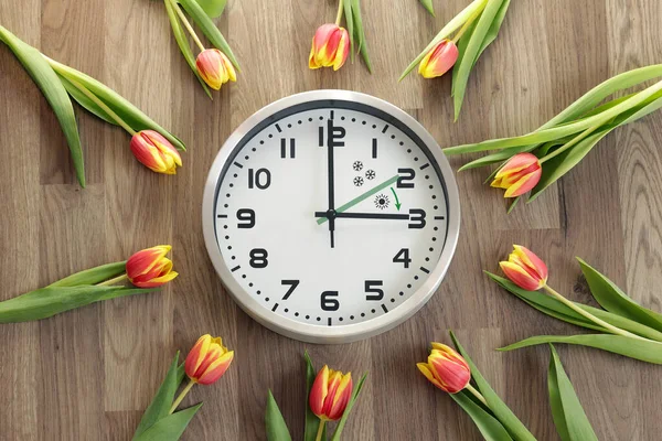 A clock shows three hours. Tulips are lying around. A symbol of the change of time. Daylight saving time. Moving the hands forward. The green arrow indicates the direction of movement of the hands.